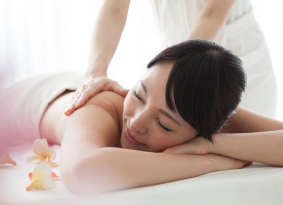 A woman lying on a massage table receiving a shoulder massage, with frangipani flowers nearby.