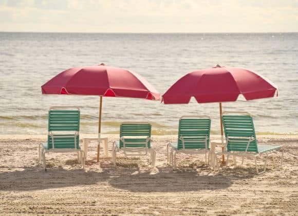 Four beach chairs arranged in pairs under two pink umbrellas on a sandy beach.
