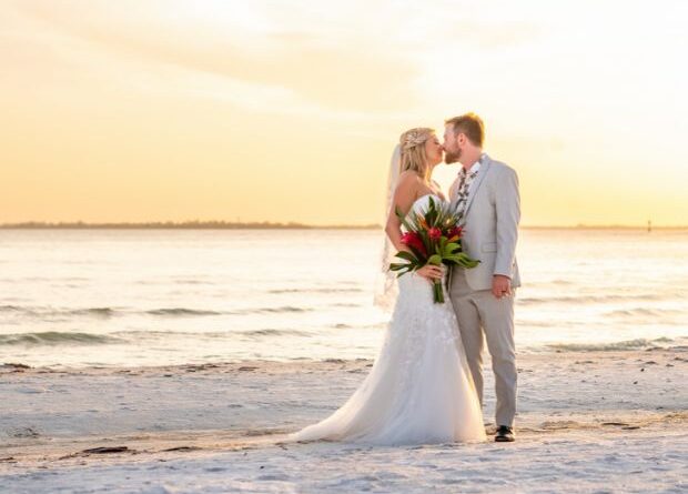 A bride and groom kissing on a beach at sunset, the bride holding a tropical bouquet.