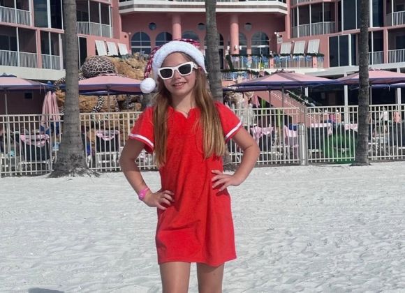 A young girl in a red dress, white sunglasses, and a Santa hat standing on a beach in front of a pink resort.