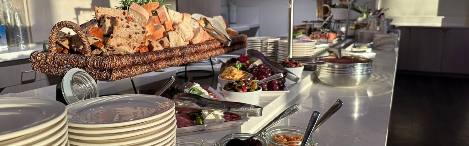 A buffet table with assorted breads, plates, cheeses, fruits, and spreads arranged neatly in a well-lit room.